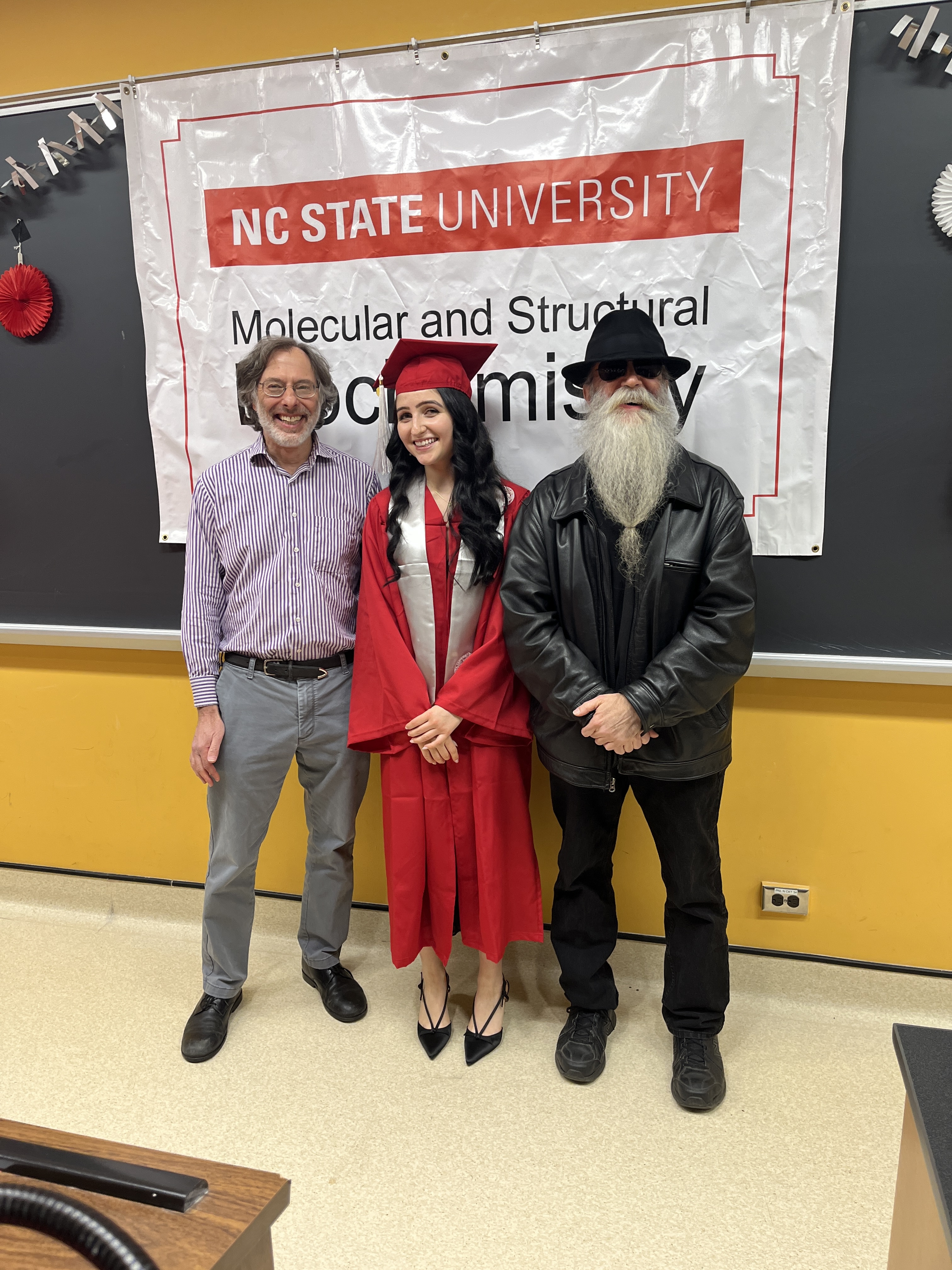 Molly at graduation in cap and gown, standing with two of her professors in front of an NC State Molecular and Structural Biochemistry banner